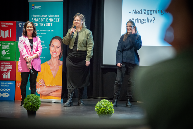 Tre kvinnor på scen. Kvinnan in mitten, Jennie-Lie Wickström pratar i en mikrofon. Kvinnan till höger i bild vinkar. Kvinnan vänter i bild har en rosa kavaj på sig. 