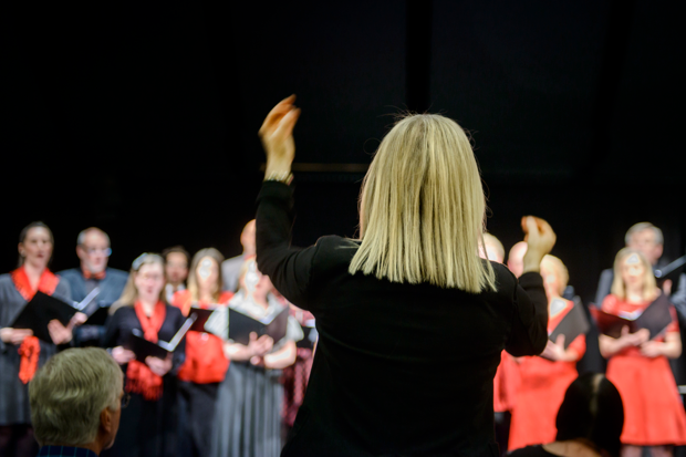 En körledare med ryggen mot kameran dirigerar en kör. I bakgrunden står flera körsångare klädda i svart och rött och sjunger från noter.