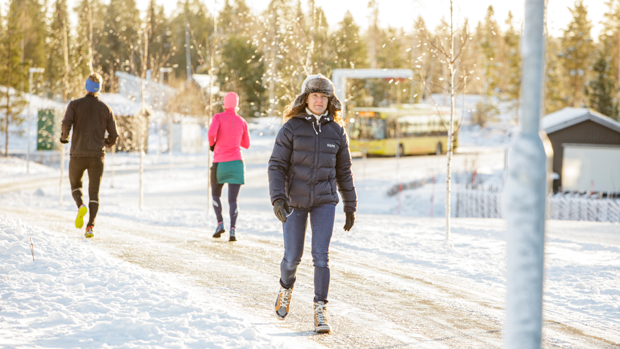 Vinterklädd kvinna promenerar. Två personer joggar bort från kameran. En elbuss laddar i bakgrunden.