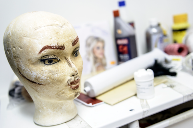 En vit frigolitdocka med handritade ögonbryn, ögon, näsa och mun står på ett bord i en ateljé eller verkstad. Runtom syns diverse konst- och hobbyartiklar som sprayflaskor, färgtuber, en ritning av ett ansikte och andra material.