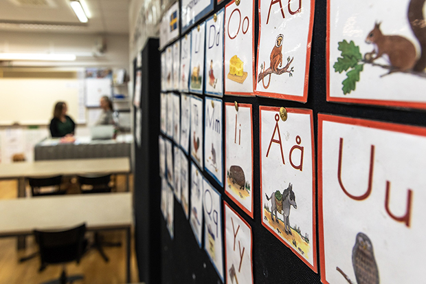 En klassrumsvägg med alfabetet uppsatt, där varje bokstav är illustrerad med en bild, exempelvis en ekorre för bokstaven 'E'. I bakgrunden syns ett klassrum med suddiga konturer av två personer som samtalar vid en kateder.