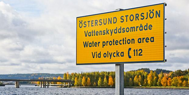 Gul skylt med text om att det är ett vattenskyddsområde. I bakgrunden syns vatten, en bro och träd i höstfärger.
