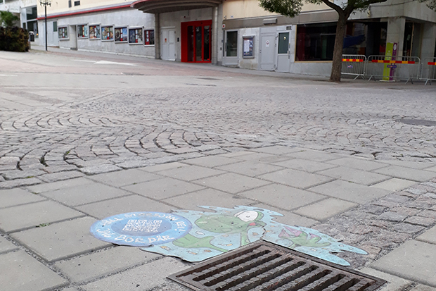 Bild på gatubrunn på en kullerstensväg i centrala Östersund. Vid brunnen ligger en dekal med texten Håll Storsjön ren.