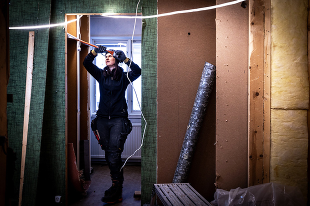 En kvinna i arbetskläder arbetar med elinstallationer i en byggarbetsplats. Hon står i en dörröppning och drar en elkabel, omgiven av isolering, verktyg och byggmaterial.