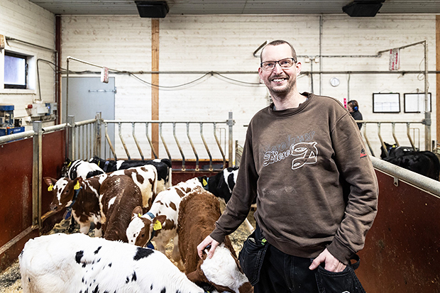 En man i arbetskläder står och ler framför en grupp kalvar i en ladugård. Kalvarna är i en inhägnad med halmbädd, och bakom dem syns en ljus trävägg och stallinredning.