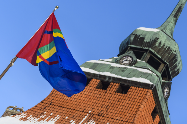 Rådhusets torn med snö på och en blå himmel i bakgrunden. Framför tornet syns en samisk flagga.