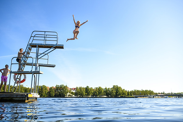 Ung flicka hoppar från ett hopptorn vid en sjö. Bakom henne är fler barn på väg uppför stegen.