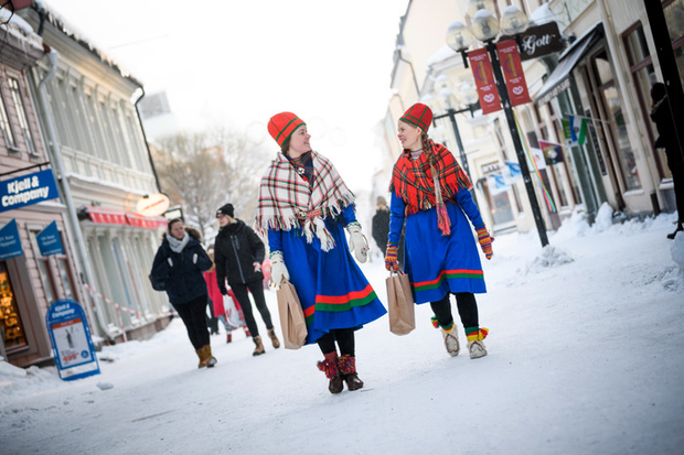  Två kvinnor i samisk folkdräkt. De har båda blåa klänningar med en röd och grön rand nertill och röd hög huvudbonad. Ena kvinnan har en vit och rödrutig sjal över axlarna och den andra har en röd och grönrutig sjal.