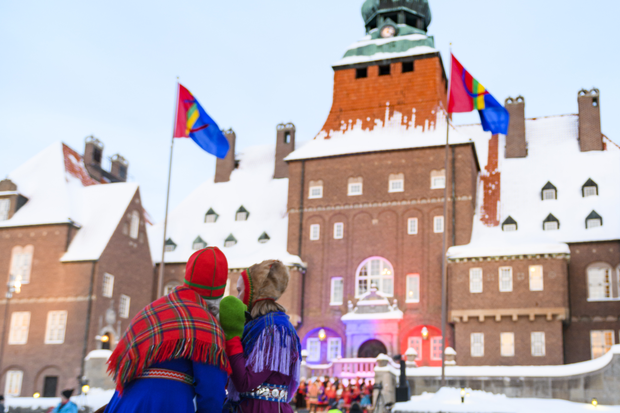I förgrunden två personer i samisk dräkt. den ena har en rödrutig sjal över axlarna och en blå klänning samt röd mössa. Den andra personen har en lila klänning med en blå sjal och en lurvig mössa. I bakgrunden Rådhuset med två samiska flaggor på flaggstänger framför.