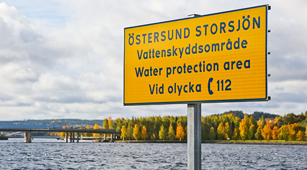En skylt i förgrunden med texten ÖSTERSUND STORSJÖN Vattenskyddsområde Water protection area Vid olika, ring 112, och Storsjön och Frösön i höstfärger i bakgrunden.