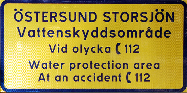 En gul skylt med texten ÖSTERSUND STORSJÖN Vattenskyddsområde. Vid olycka ring 112. Water protection area At an accident, call 112