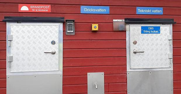 Två metalluckor med dörrhandtag. Ovanför skyltar med texten Brandpost, dricksvatten och tekniskt vatten. Mellan luckorna finns en dosa för portkod och en knapp. Under knappen finns en tappkran.
