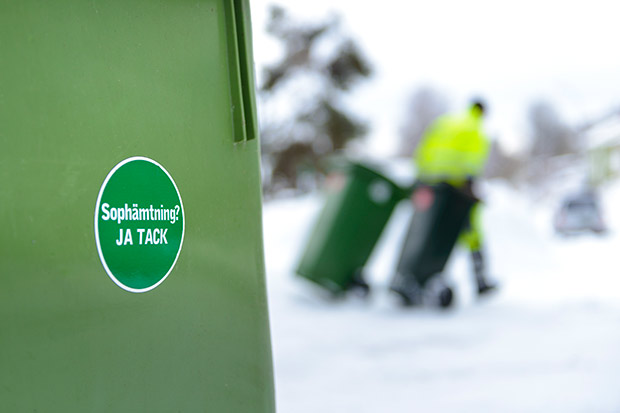 Sopkärl med grön rund dekal. I bakgrunden en person med varselkläder som drar två sopkärl