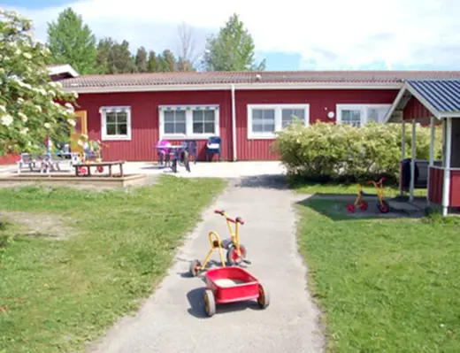 en röd byggnad i bakgrunden, i förgrunden syns en trehjuling, en lekstuga, sandlåda och gräsytor