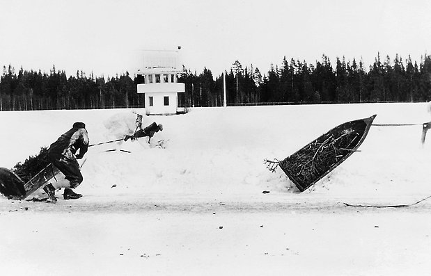 Svartvitt foto på ren med släde som hoppat upp på en snövall samt en person som lyfter släden.