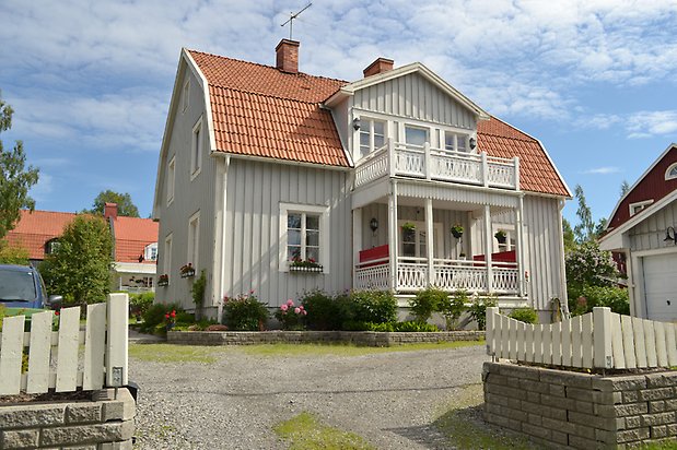 Forellen 8, Villa i grå träpanel, vita fönster med spröjs och balkong och förstukvist med snickarglädje.