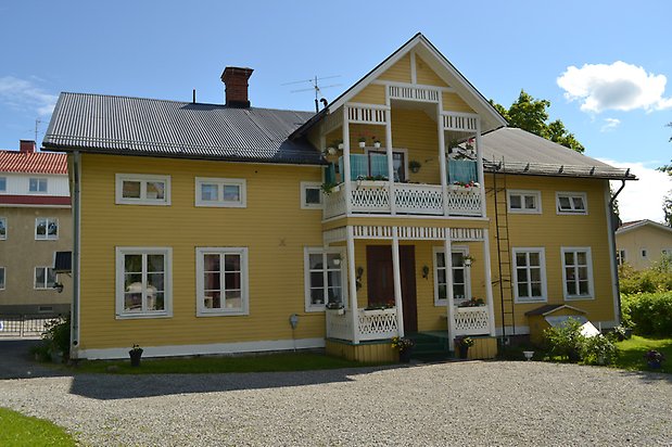 Konvaljen 8, Villa med gul, liggande träpanel och vita fönster med spröjs, förstukvist och balkong med snickarglädje.