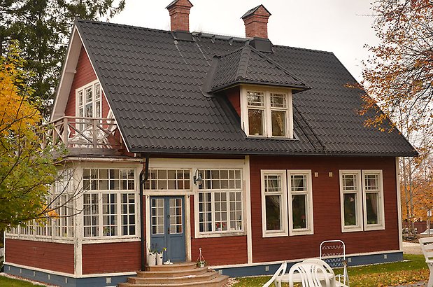 Alnmåttet 4. Villa i röd, liggande panel, veranda med många fönster med spröjs, blå dubbeldörrar. Svart tak och takkupa.