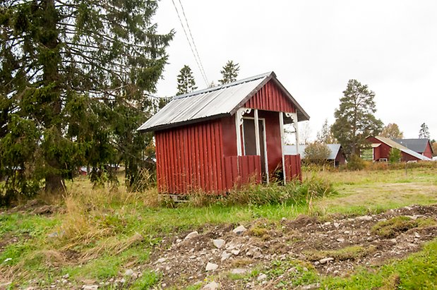 Korsta 2:4. Liten byggnad i röd panel med förstukvist med svart dörr.