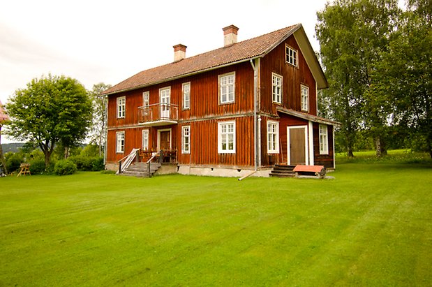 Digernäs 2:7. Hus i två våningar i röd panel med balkong i metall, lite tillbyggnad med brun dörr och fönster med vita spröjs.