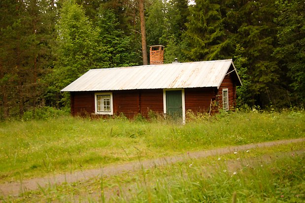 Orrviken 2:12. Timrad, brun stuga med grön dörr och fönster med vita spröjs.