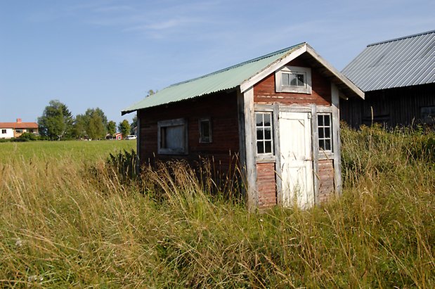 Norderöns-Backen 1:12. Liten byggnad i röd liggande panel, vit dörr med fönster med spröjs.