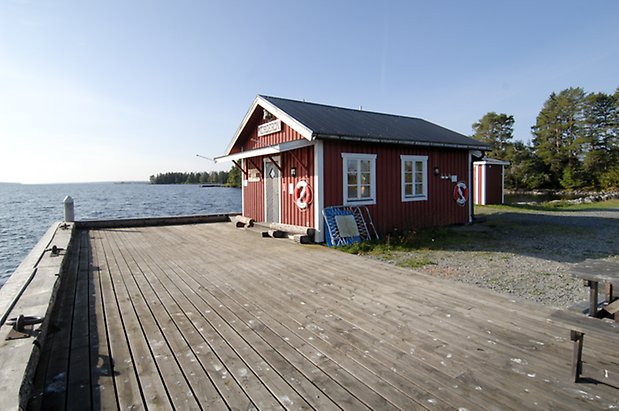 Önsta 1:36. Byggnad i röd panel med grå dörr och fönster med vita spröjs, tak över ingången, livboj på väggen. Brygga och vatten.