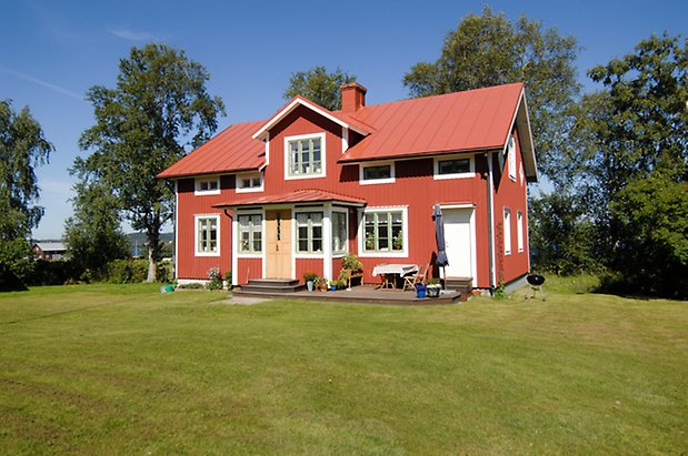 Namn 2:18. Villa i två våningar i röd panel med vita detaljer, fönster med gröna spröjs, takkupa med fönster, punschveranda med gul dörr. 