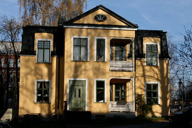 Stenbyggnad i tre våningar i gul puts med vita detaljer. Gröna fönster med spröjs, gröna dörrar och svart plåttak.