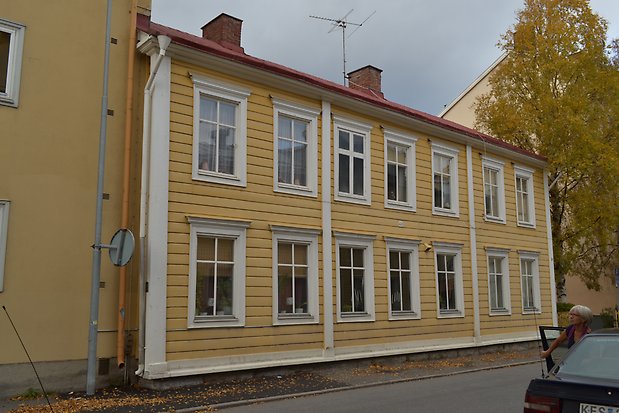Träbyggnad i två våningar,  i gul, liggande panel, vita knutar och fönsterfoder och fönster med vita spröjs. 