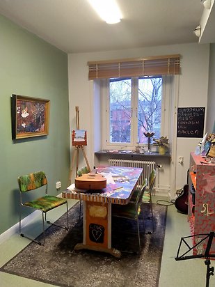 Rum i ateljé med ett bord med en gitarr på bordet. Tavlor på väggar och ett fönster mitt i bilden.