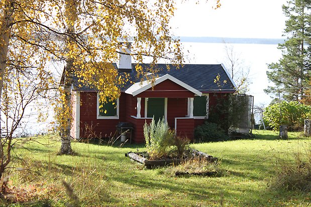 Kattfoten 1.  Hus i röd, liggande panel, vita fönster med gröna luckor, grön dörr, och svart tak.