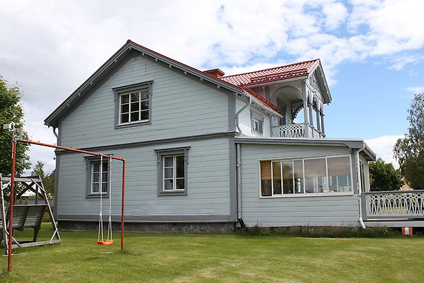 Orrviken 5:1. Villa i vit liggande panel med grå fönster med vita spröjs. Punschveranda och balkong med grå snickarglädje. Rött tak.