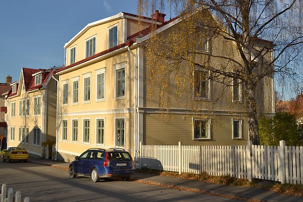 Trähus i tre våningar i ljusgul, liggande panel med vita detaljer och gröna fönster med spröjs. Stengrund och rött plåttak.