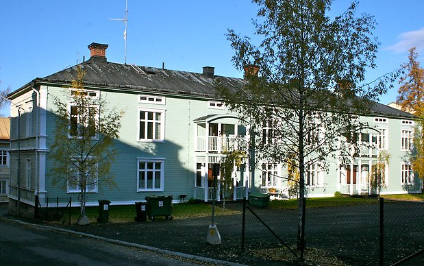 Träbyggnad i två våningar i ljusgrön, liggande panel med vita detaljer, vita fönster med spröjs, svart plåttak. Två förstukvistar med balkong, 