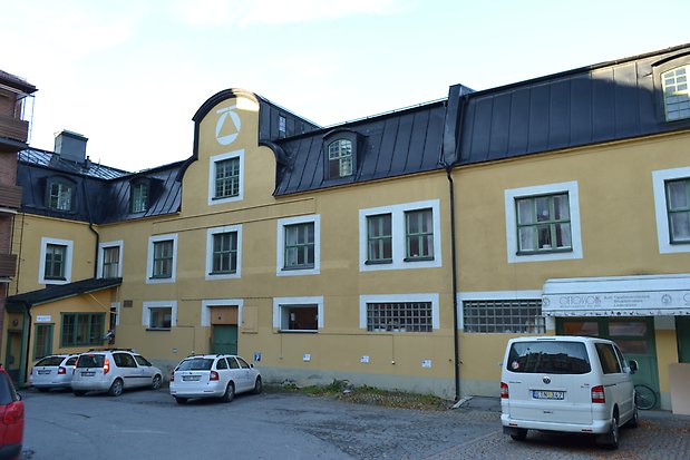 Stenbyggnad i tre våningar i gul puts med vita detaljer, gröna fönster med spröjs, svart plåttak med takkupor.